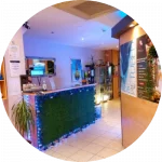 Reception area with a blue mosaic-patterned desk, tiled floor, various plants, and posters on the wall, creating a warm and welcoming ambiance for tanning.