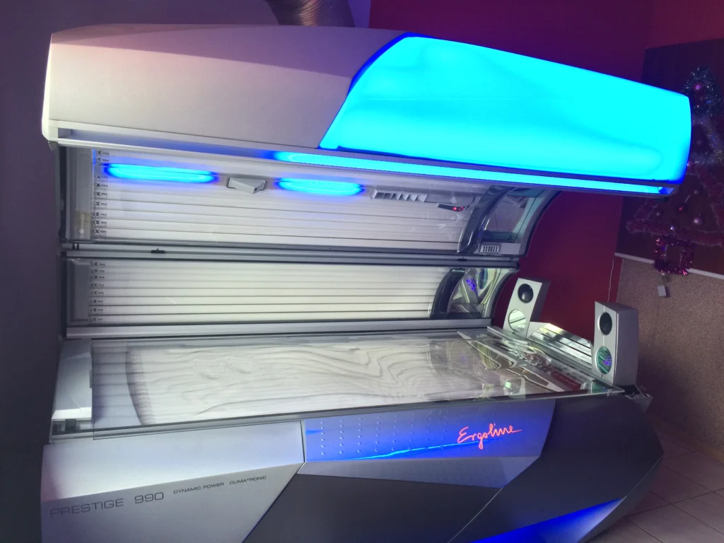 An Ergoline Prestige 990 tanning bed is open and ready for use. The top canopy of the bed has blue accent lighting and houses several fluorescent tube lights designed for tanning.